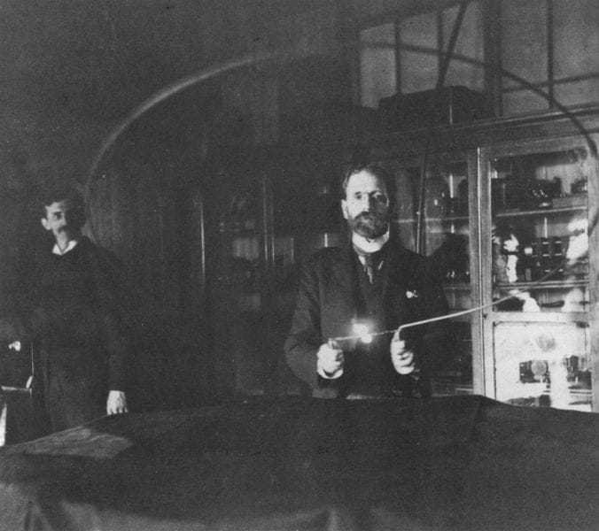 Robert Underwood Johnson holding a loop with an incandescent lamp. Tesla is in the background at the switch.