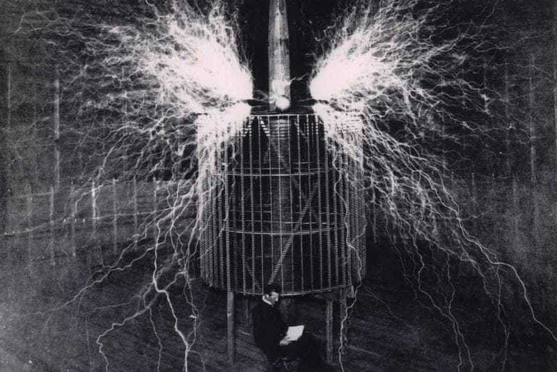 Burning the nitrogen of the atmosphere. This result is produced by the discharge of an electrical oscillator giving twelve million volts. The electrical pressure, alternating one hundred thousand times per second, excites the normally inert nitrogen causing it to combine with oxygen. The flame-like discharge measures sixty-five feet across. Tesla sits in front of the oscillator in a second exposure.
